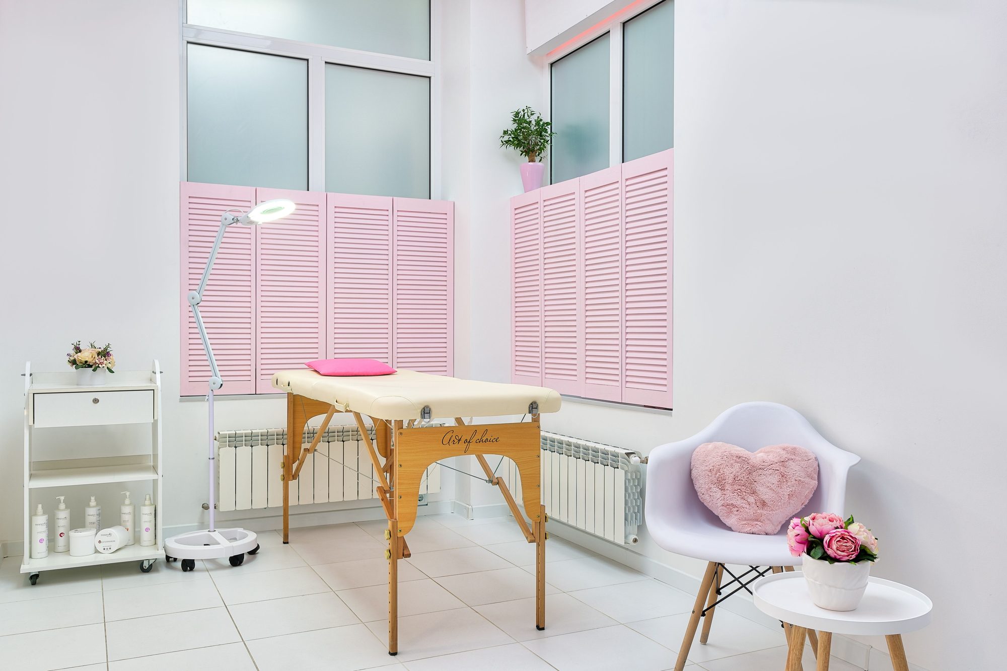 Sunlit spa treatment room at Verbena & Clay with a cushioned treatment table, soft pink window shutters, and fresh flowers